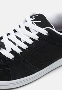 Sorte semskete sneakers har hvite lisser, perforerte detaljer og en gummisåle med en kontrastfarget hvit stripe rundt bunnen.