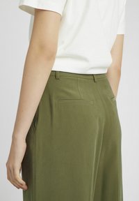 Pantalons verts kaki avec une texture lisse, présentant des poches arrière soignées et une coupe ajustée, associés à unsimple t-shirt blanc.