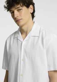 Bílá košile s krátkým rukávem a knoflíkovým zapínáním vyrobená z lehkého materiálu, s volným střihem, notched límcem a jemnou texturou.