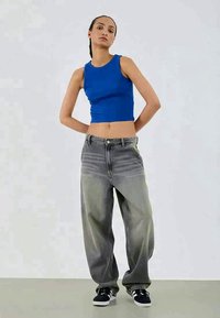 Γυναίκα που στέκεται με τα χέρια πίσω από την πλάτη, φορώντας μπλε αμάνικο τοπ, φαρδιά ξεθωριασμένα γκρίζα τζιν και μαύρα αθλητικά παπούτσια.