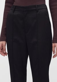 Femme portant un pantalon noir ajusté et un top côtelé à manches longues brun foncé, mains relâchées le long du corps sur un fond uni.