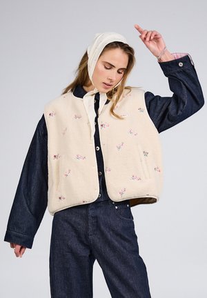 Mujer con un chaleco de forro polar crema con flores sobre una chaqueta y pantalones de mezclilla oscuros, con un pañuelo claro en la cabeza, levantando un brazo contra un fondo liso.