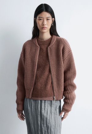 Strikket, oversized bomberjakke i støvet rosafarve med ribbet tekstur; lynlåslukning og tætsiddende manchetter. Båret over en matchende, let sweater.