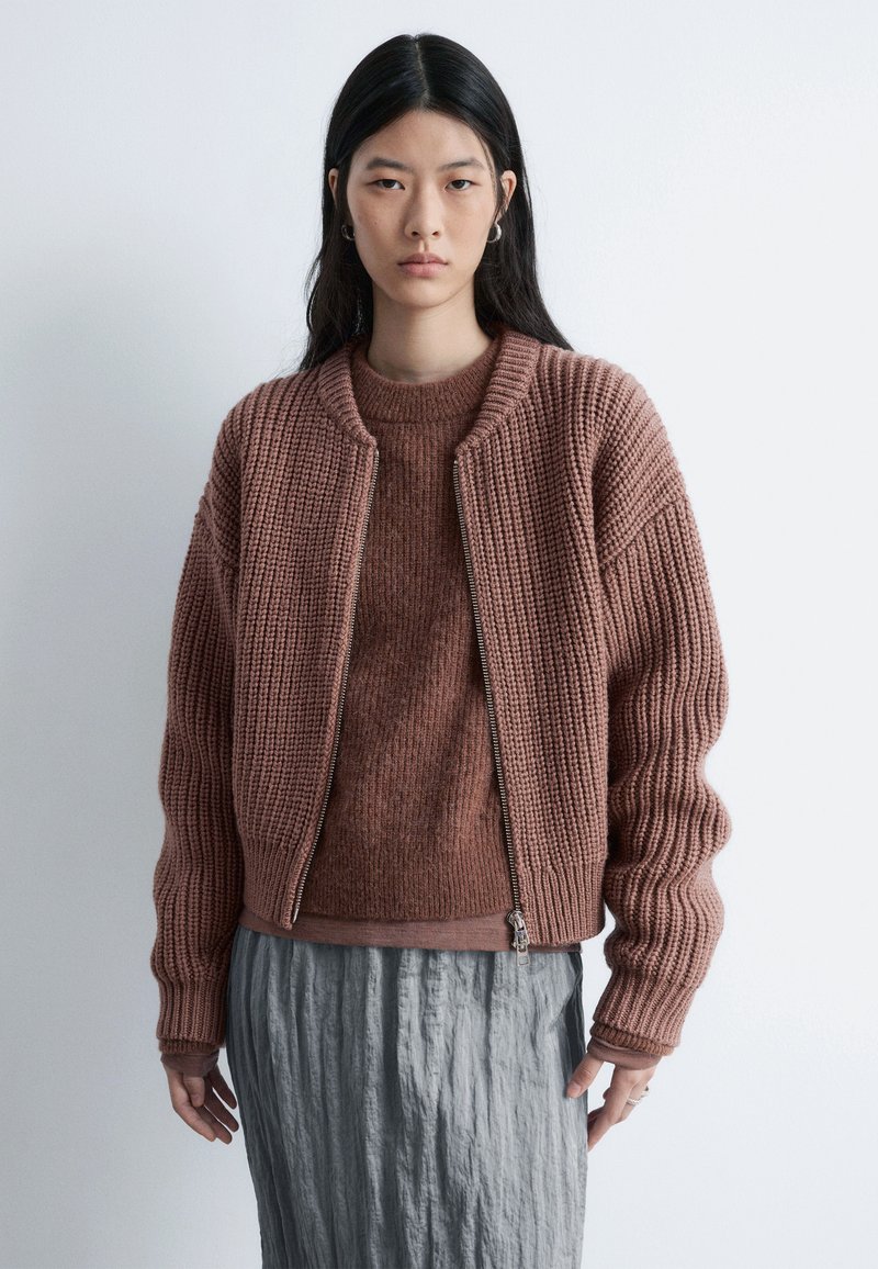 Gestrickte, übergroße Bomberjacke in staubigem Rosa mit geripptem Muster; Reißverschluss und eng anliegende Bündchen. Drüber getragen ein passender, leichter Pullover.