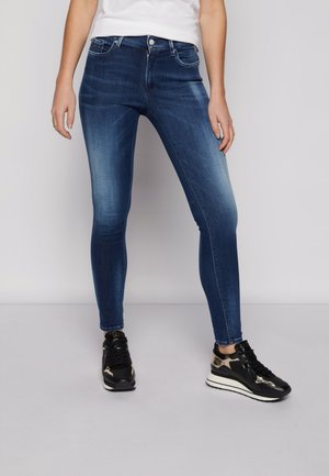 Persona con vaqueros pitillo azul deslavado combinados con zapatillas plataforma negras y doradas, de pie contra un fondo liso.