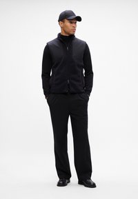 Colete de felpa preto sobre um suéter de gola alta preto, combinado com calças pretas e sapatos pretos. O modelo usa um boné preto combinando.