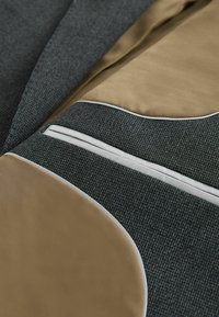 Tissu texturé gris avec des accents beiges, présentant un intérieur lisse en satin, des garnitures blanches et un détail de poche sur mesure.