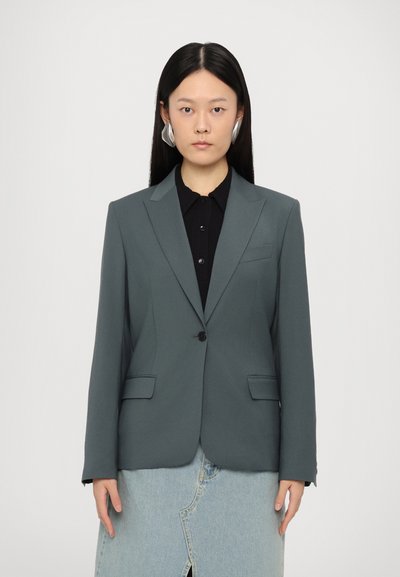 Blazer gris ajusté avec revers crantés, fermeture à un bouton et deux poches avant. Porté par-dessus une chemise noire à col et une jupe en denim.