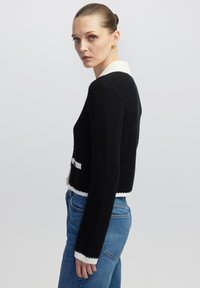 Schwarzer strukturierter Pullover mit einem weißen Kragen und Besatz, kurzes Design, lange Ärmel und einem dekorativen Schleifenakzent in der Taille.