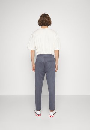 Person mit lockigem Haar steht mit dem Rücken zur Kamera, trägt ein weißes kurzärmeliges Hemd, blau-graue Hose und weiße Turnschuhe mit rotem Schriftzug.