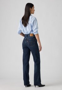 Sieviete stāv sāniski, ģērbta zila svītrainā krekla, kas ielikts augstās jostasvietas tumši zilajās džinsās un melnās papēžu kurpēs uz vienkārša fona.