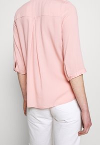Light pink, textured blouse with three-quarter sleeve and button detail; back pleat design; paired with white pants.