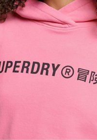 Růžová mikina s kapucí a jemnou texturou, na přední straně se výrazným černým logem "SUPERDRY" a symbolem registrované ochranné známky.