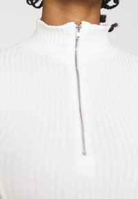 Witte geribde top met ritssluiting en een hoge kraag, voorzien van een zilverkleurige ritssluiting aan de voorkant, gemaakt van een gestructureerde gebreide stof.