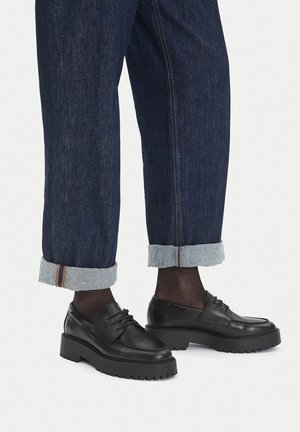 Schwarze Lederschuhe mit einer dicken, geriffelten Sohle und Schnürdesign, kombiniert mit hochgekrempelten, dunkelblauen Jeans aus Denim mit kontrastierender Naht.