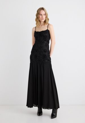 Frau mit blonden, welligen Haaren, die ein schwarzes, eng anliegendes Kleid mit floralem Muster, dünnen Trägern und einem fließenden Rock trägt, kombiniert mit schwarzen Stiefeletten.