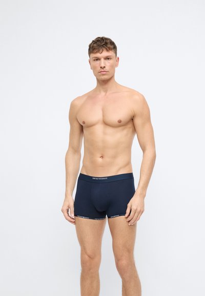 Tumši zili boxer šortai ar pieguļošu dizainu, elastīgu jostu ar zīmola logo un gludu auduma tekstūru. Modelis ir bez krekla.