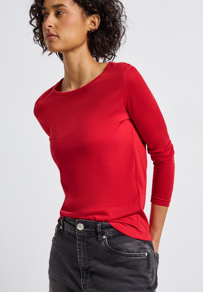 Frau mit lockigem Haar, die ein enges rotes Langarmshirt trägt, das in hoch geschnittene, ausgebleichte schwarze Jeans gesteckt ist, mit Händen hinter dem Rücken.