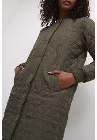 Femme aux cheveux bouclés portant un long manteau matelassé vert olive avec des boutons-pression et les mains dans les poches avant sur fond uni.