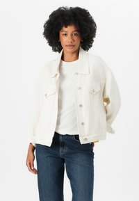 Hvit denimjakke med en avslappet passform, stor krage, sølvknapper og to frontlommer, parret med en hvit t-skjorte og mørke jeans.