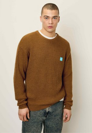 Brauner Strickpullover mit gerippter Textur, runder Ausschnitt und lockerer Passform. Mit einem blauen Markenpatch auf der linken Seite. Wird über einem weißen Shirt getragen.