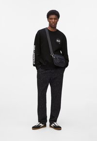 Mann trägt einen schwarzen Pullover mit Logo und weißen Texten auf dem Ärmel, gemusterte schwarze Hose, schwarz-weiße Sneakers und eine schwarze Umhängetasche.