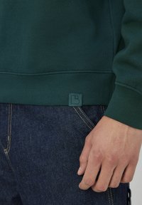 Sweatshirt vert avec ourlet et poignets côtelés, arborant une étiquette de logo texturée. Accompagné d'un jean en denim bleu foncé, montrant des coutures contrastées.