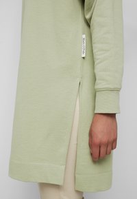 Top verde chiaro a maniche lunghe con spacchi laterali, polsini a costine e un'etichetta tessuta sull'orlo. Realizzato in un tessuto morbido con una consistenza liscia.