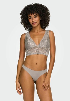 Frau mit lockigem Haar, die ein graues Spitzen-Bralette und passende nahtlose Unterwäsche trägt, steht vor einem schlichten hellen Hintergrund.