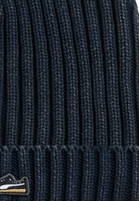 Gorro de punto acanalado en color navy con acabado texturizado y un pequeño parche con el logo en la parte frontal, que presenta detalles de costura en contraste.