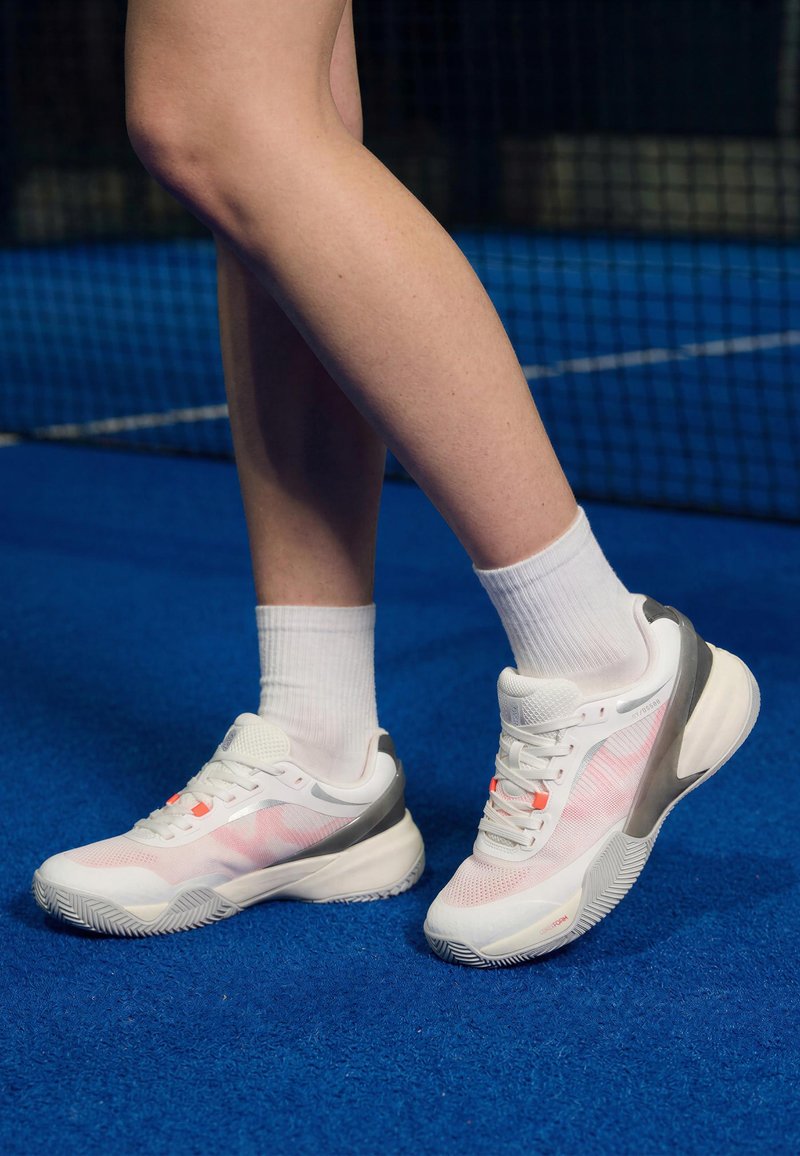 Jambes portant des chaussettes blanches et des chaussures de sport blanches et grises, debout sur un court de tennis bleu près du filet.