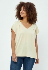Helles beige V-Ausschnitt-Oberteil mit kurzen Ärmeln, das feine Falten am Ausschnitt und einen lockeren Schnitt aufweist, kombiniert mit blauen Jeans.