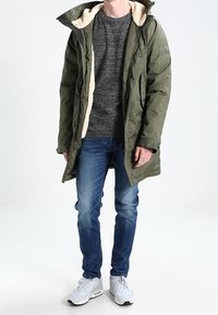 Grön vadderad parkas med en vit fleecefoder, bärs över en grå texturerad tröja, ihop med blå jeans och vita sneakers.