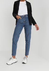 Osoba w czarnym żakiecie, białym t-shircie, niebieskich dżinsach z wysokim stanem i białych trampkach, stojąca na tle jasnego, jednolitego tła.