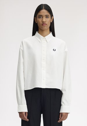 Femme aux cheveux noirs raides portant une chemise blanche ample à boutons avec un petit emblème noir brodé sur le côté gauche de la poitrine.