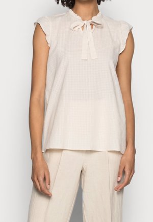 Blouse crème sans manches avec col à volants et col à nouer, associée à un pantalon large crème assorti.