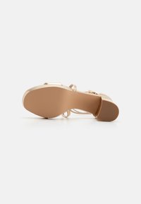 Sandal med en ljusbeige, glansig yta; snördesign; elegant klack; gummisula med texturerat grepp.