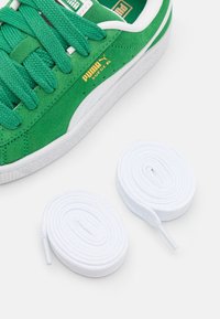 Gröna mockasneakers med vit gummisula och snören. Har texturerade snören och en guldfärgad "PUMA"-logotyp på sidan. Inkluderar två vita snören.