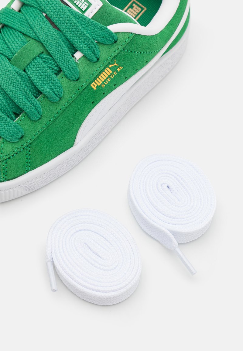 Baskets en suède vert avec semelle et lacets en caoutchouc blanc. Elles présentent des lacets texturés et un logo "PUMA" doré sur le côté. Comprend deux lacets blancs.