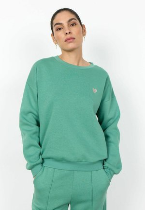 Frau mit dunklen, zurückgebundenen Haaren, die einen grünen Pullover und passende Hosen trägt, mit den Händen in den Taschen, vor weißem Hintergrund.