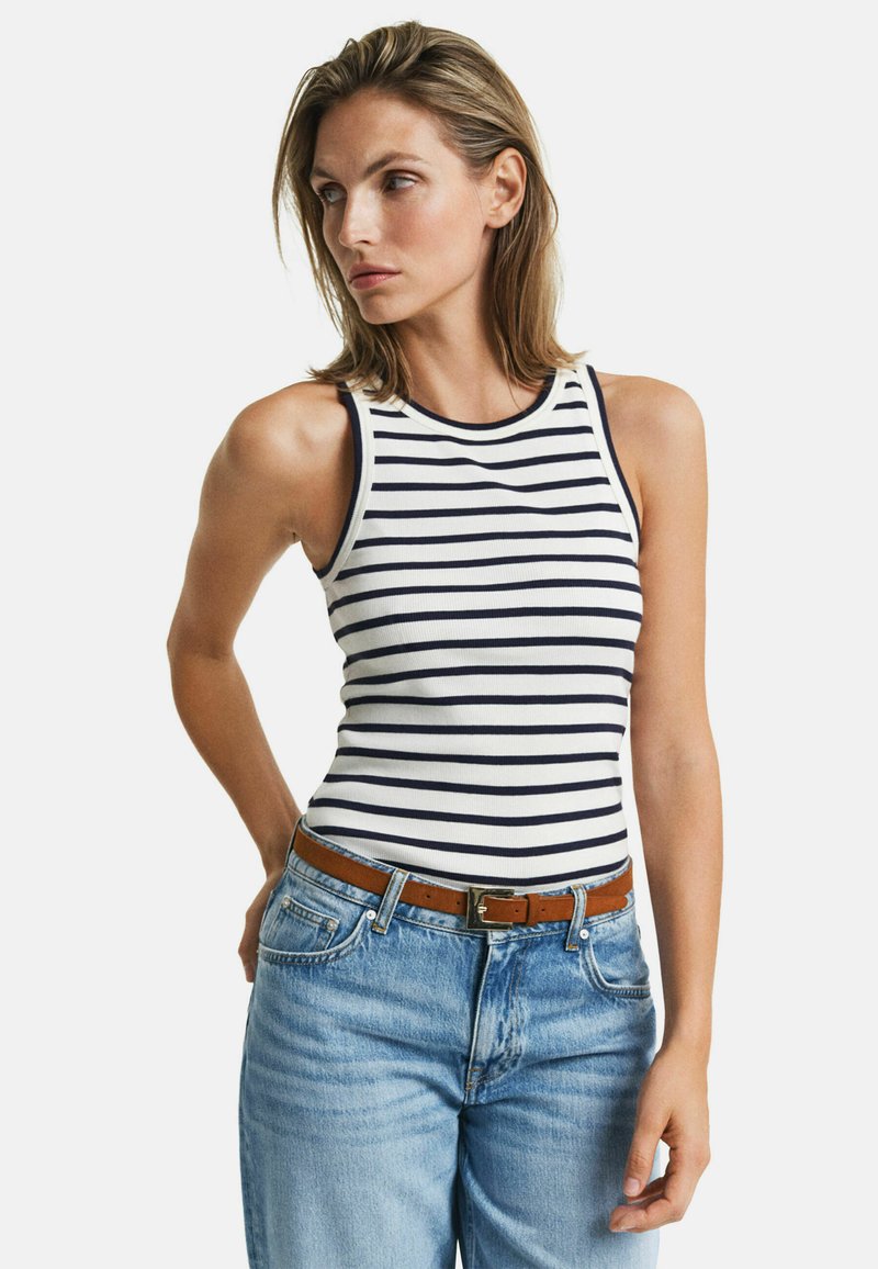 Camiseta de tirantes a rayas en blanco y azul marino, con textura acanalada y diseño sin mangas. Combinada con jeans azules y un cinturón marrón.