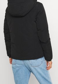 Schwarze, puffy Jacke mit Kapuze. Mit elastischen Bündchen und geradem Schnitt. Kombiniert mit hellblauen Jeans, die ein lässiges Outfit zeigen.