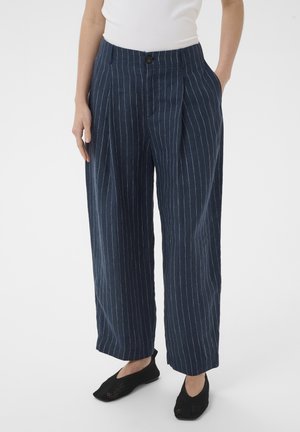 Femme portant un pantalon large bleu marine à fines rayures blanches verticales, un haut blanc sans manches et des chaussures noires à enfiler.