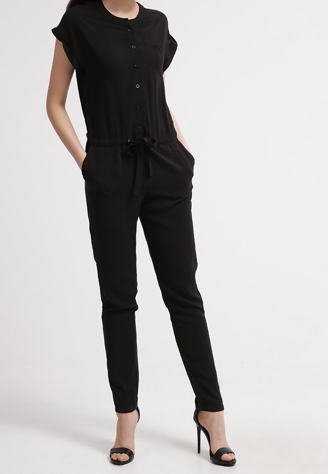 Femme portant une combinaison noire boutonnée à manches courtes, taille avec cordon, mains dans les poches et sandales noires à talons hauts.