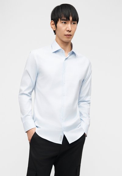 Άνδρας με μαύρα μαλλιά, ντυμένος με ανοιχτό μπλε πουκάμισο με κουμπιά και μαύρο παντελόνι, στέκεται με τα χέρια στις τσέπες πάνω σε απλό φόντο.