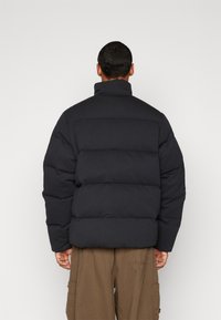 Jachetă puffer neagră cu guler înalt și design cu matlasare orizontală. Prezintă o croială relaxată, cu mâneci largi și o textură netedă.