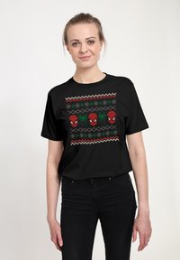 Černé bavlněné tričko má grafický design s opakujícími se tvářemi Spider-Mana, zelenými akcenty a cik-cak vzorem, umístěným proti svátečnímu pozadí.
