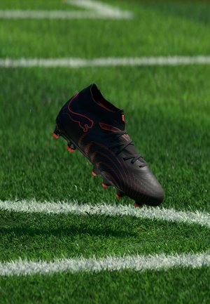 Sort og rød fodboldstøvle svæver over hvide markeringer på grøn græs, vist fra en sidevinkel med synligt mærkelogo.