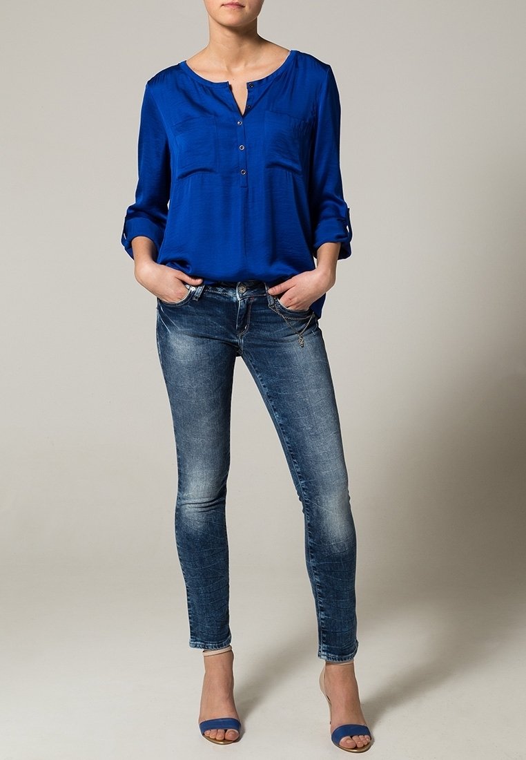 Blå silkblus med knappfront och uppvikta ärmar, parat med slitna blå denimjeans och blå klacksandaler.