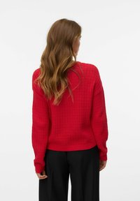 Femme avec de longs cheveux ondulés portant un pull rouge texturé et un pantalon noir, de dos contre un fond blanc uni.